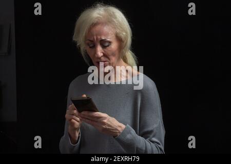 Ritratto classico di una donna bionda anziana in Studio su sfondo nero. Legge un messaggio sul telefono Foto Stock