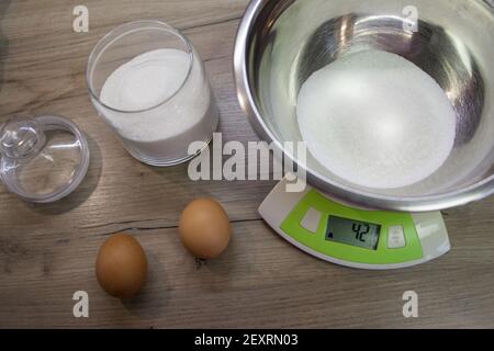 Zucchero bianco in una ciotola sulla bilancia e due uova sullo sfondo del legno. Il secondo passo per la preparazione di muffin, torta, biscotto. Cottura passo dopo passo Foto Stock