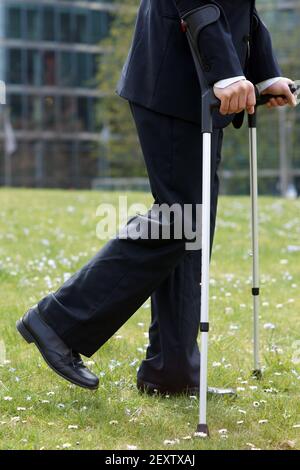 Uomo d'affari cammina attraverso un parco in città con le stampelle dopo lesioni alle ginocchia Foto Stock