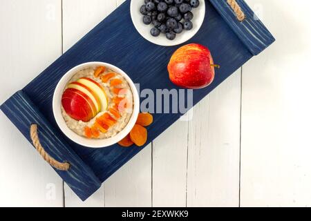 vista dall'alto sul recipiente con porridge e farina di avena con mirtilli e fette di mela. deliziosa colazione salutare. servita su vassoio blu e tavolo di legno bianco Foto Stock