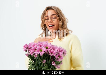 Sorpresa bella giovane donna riceve bouquet di fiori come regalo sorridente li guarda mentre si sta in piedi su sfondo bianco studio. Foto Stock