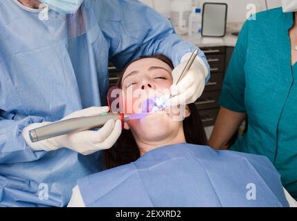 dentista e infermiera che lavorano con dispositivi di protezione individuale, paziente donna sulla sedia, denti al neon leggeri sbiancanti. Durante la pandemia del coronavirus Foto Stock