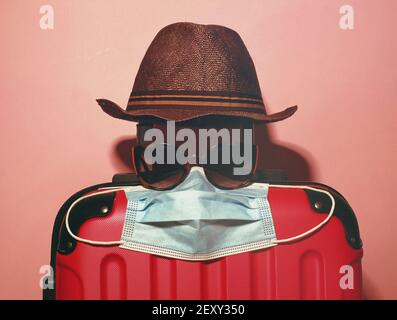 Valigia, cappello estivo, occhiali da sole e maschera facciale su sfondo rosa pastello. Concetto di vacanza estiva sotto le restrizioni del coronavirus. Mare sabbia sole. Foto Stock