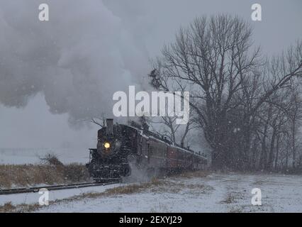 Vista di una locomotiva a vapore antica restaurata che soffia fumo e. A vapore che viaggia attraverso le terre e la campagna in una tempesta di neve Foto Stock