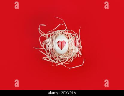 Uovo bianco con forma a cuore in nido decorativo su sfondo rosso. Foto Stock