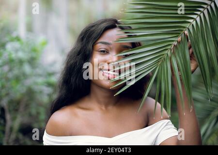 Ritratto tropicale di moda. Giovane attraente modello donna skinned scuro in camicia bianca, tenendo in mano foglia verde di palma mentre posa in bella Foto Stock