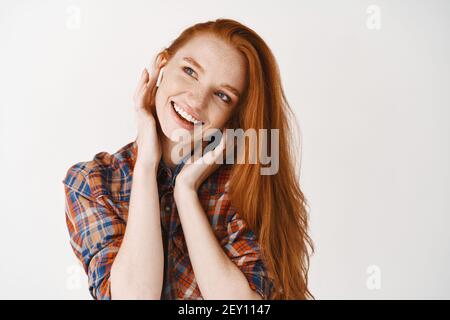 Caucasica ragazza con i capelli zenzero e pelle pallida ascoltare musica in cuffie wireless, sorridente e guardando in alto a sinistra, in piedi su bianco Foto Stock