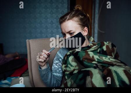 una ragazza malata si siede avvolta in una coperta con una maschera sul viso. misurare la temperatura con un termometro durante un'epidemia. cattiva salute Foto Stock