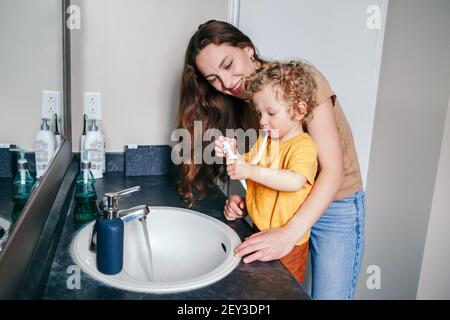 Giovane madre caucasica che aiuta il bimbo a spazzolare i denti in bagno a casa. Igiene sanitaria e routine mattutina per bambini. Mamma che aiuta a sostenere Foto Stock