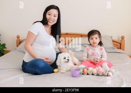 Buona Pasqua. Madre cinese asiatica incinta con la bambina che gioca con le uova di Pasqua colorate sul letto a casa. Bambino e genitore festeggiando la traditio Foto Stock