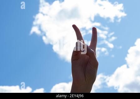 Mano con due dita in alto nel simbolo della pace o della vittoria, un segno per la lettera V in lingua dei segni. Isolato su sfondo blu cielo con nuvole, COP Foto Stock