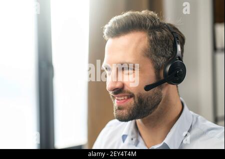 Bell'uomo europeo dai capelli marrone scuro, indossando cuffie, guardando lontano, lavorando come operatore, sorridente amichevole, sicuro, indossare una camicia blu formale, sul posto di lavoro, amore per aiutare le persone Foto Stock