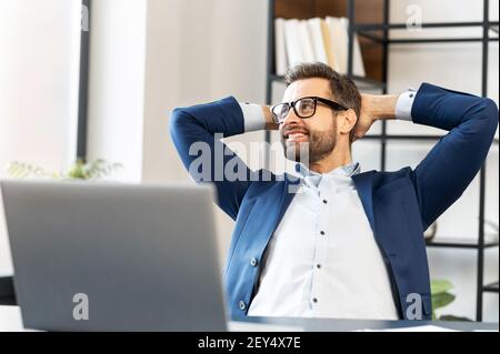 Uomo sorridente con gli occhiali seduti al computer portatile, tenendo le mani dietro la testa, rilassante, guardando il bel tempo fuori dalla finestra, lavora in un ufficio confortevole, ha fatto il lavoro in modo produttivo e ha ottenuto elogi Foto Stock