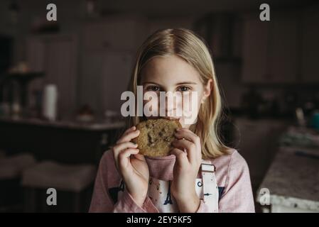 Tween ragazza che mangia un grande biscotto di chip di cioccolato in cucina con grembiule Foto Stock