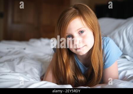 Giovane ragazza dagli occhi blu con capelli rossi felice che si posa sul letto bianco. Foto Stock