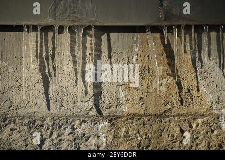 ghiaccioli congelati trasparenti e appuntiti appesi alle ombre della fusione industriale di tubi su cemento grungo parete esterna dell'edificio sfondo orizzontale Foto Stock