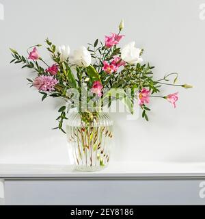 Un bouquet minimalista di tulipani bianchi, eustoma rosa, giacinto, eucalipto in vaso di vetro scanalato su un pannello bianco di un camino artificiale. Quadrato Foto Stock