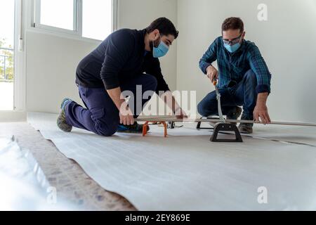 Due giovani uomini con maschera tagliando un laminato di legno con una ghigliottina per installare un pavimento in legno nel loro appartamento Foto Stock
