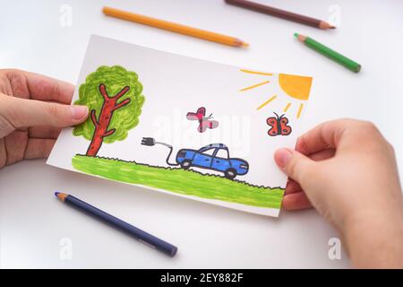 Ragazzo giovane che tiene un disegno chillike di un'automobile elettrica che va per una guida all'aperto nelle sue mani. Primo piano. Foto Stock