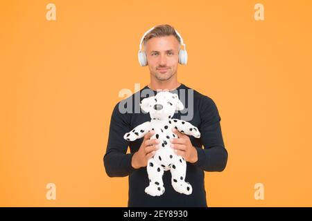 Bell'uomo di mezza età tenere morbido cane giocattolo per giocare ascoltando la musica in cuffia sfondo giallo, adozione. Foto Stock