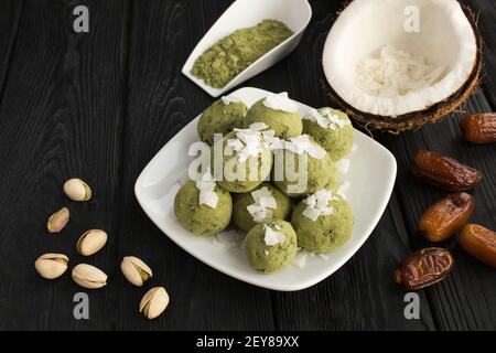 Palle energetiche con polvere di matcha, pistacchi, datteri e patatine di cocco nella piastra bianca sullo sfondo di legno nero. Primo piano. Foto Stock