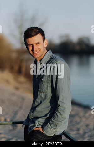 Uomo sorridente in camicia jeans in piedi fuori con le mani in tasca. Ritratto dell'uomo Foto Stock