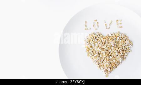 oat flakes in una forma di colpo di cuore dall'alto su un piatto bianco su sfondo bianco cibo vegetariano biologico. Foto Stock