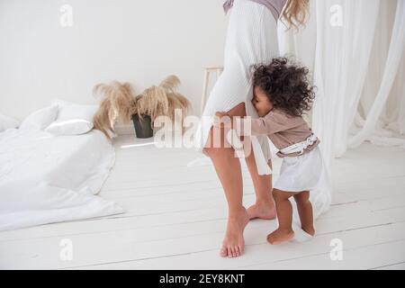 Bambina afroamericana dai capelli ricci, che si nasconde sotto l'abito della madre, sbirciando tra le gambe, divertendosi, ridendo, mostrando la lingua. Interno minimalista Foto Stock