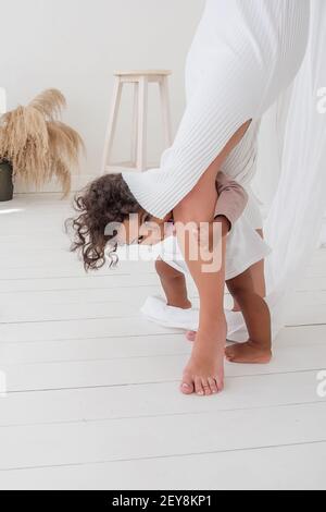 Bambina afroamericana dai capelli ricci, che si nasconde sotto l'abito della madre, sbirciando tra le gambe, divertendosi, ridendo, mostrando la lingua. Interno minimalista Foto Stock