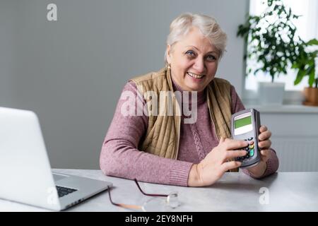 donna anziana seduta con computer portatile e terminale pos durante la quarantena Foto Stock