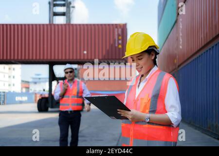 Donna lavoratrice asiatica sta controllando il contenitore dopo la riparazione completo di caposquadra che utilizza walkie talkie per comandare il sollevamento della gru contenitore in backgrou Foto Stock