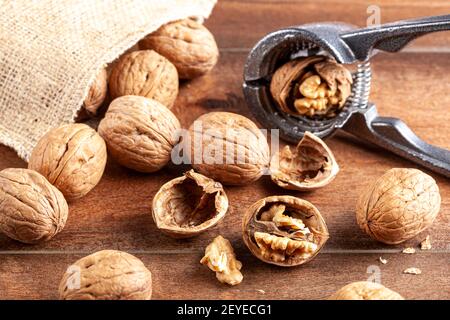 Immagine di messa a fuoco selettiva angolata delle noci in guscio sparse da un sacco vintage burlap su un tavolo di legno. Alcune sono incrinate con pinze in metallo noce Foto Stock