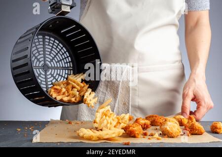 Una donna sta scaricando le patatine fritte di cialda di patate fresche dal cestino su un piano di lavoro insieme ai nocciole di pollo. Li ha fritti in friggitrice ad aria usando molto Foto Stock