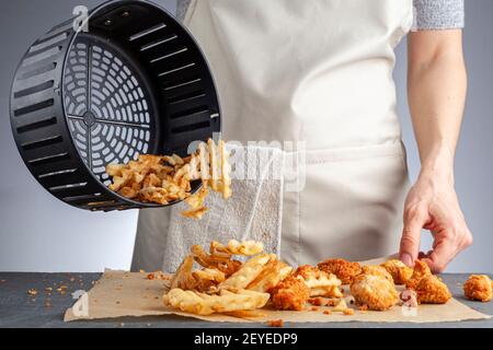 Una donna sta scaricando le patatine fritte di cialda di patate fresche dal cestino su un piano di lavoro insieme ai nocciole di pollo. Li ha fritti in friggitrice ad aria usando molto Foto Stock