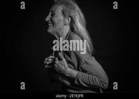 Classico ritratto drammatico in bianco e nero di una anziana bionda in Studio su sfondo nero, tiene le mani sul petto e ride Foto Stock