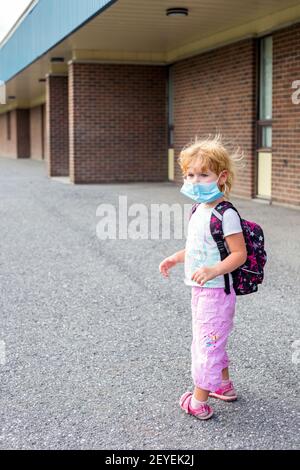 Piccolo bambino preschooler in maschera con zaino durante il virus corona vicino alla costruzione della scuola, andando a scuola o asilo. Primo giorno a scuola concetto. Foto Stock