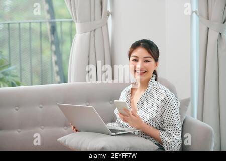 Foto di una donna asiatica incinta sorridente che usa il computer portatile e il telefono cellulare mentre si siede sul divano a casa Foto Stock