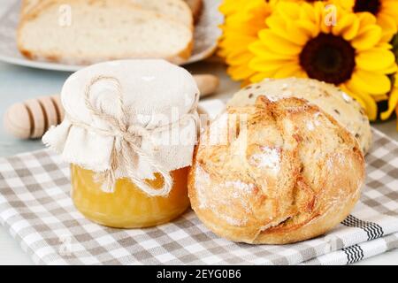 Vasetto di vetro con miele, panini e pane sul tavolo per la colazione. Bouquet di girasoli sullo sfondo. Foto Stock