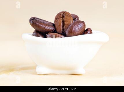 Primo piano di una piccola vasca da bagno bianca piena di chicchi di caffè tostati su uno sfondo neutro. Studio shot, messa a fuoco selettiva. Foto Stock