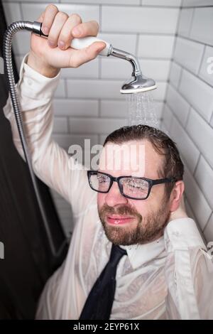 Un uomo soddisfatto e sorridente in bicchieri e vestiti si trova in bagno e versa acqua su di sé da una lattina annaffiante dopo il lavoro. Foto Stock