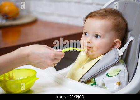 Un bambino sorridente di nove mesi si siede ad un tavolo bianco in una seggiolone e mangia con un cucchiaio da una ciotola. La mamma alimenta il bambino da un cucchiaio. Backgro sfocato Foto Stock