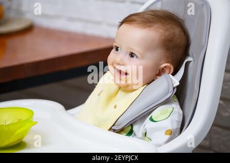 Un bambino sorridente di nove mesi si siede ad un tavolo bianco in una seggiolone e mangia con un cucchiaio da una ciotola. La mamma alimenta il bambino da un cucchiaio. Backgro sfocato Foto Stock