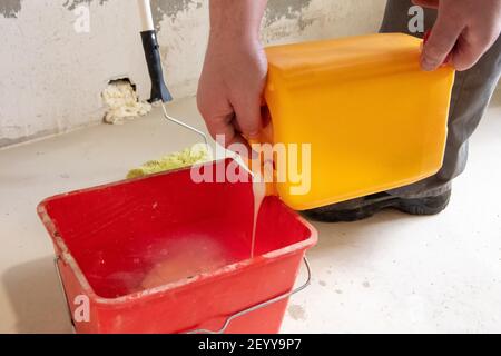 Concetto di rinnovamento. Fai da te. Mani maschili che versano vernice o colla da un contenitore in un secchio rosso, primo piano. Foto Stock