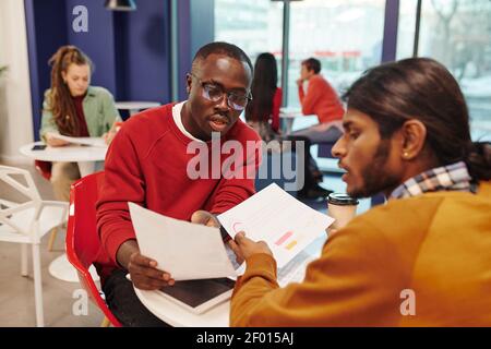 Due giovani imprenditori interculturali in casualwear guardando la carta mentre uno di loro indica i dati finanziari e li spiega al collega Foto Stock