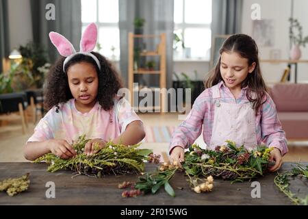 Due bambine carine di varie etnie in piedi da tavolo Davanti alla macchina fotografica e preparando le corone pasquali contro casa ambiente Foto Stock