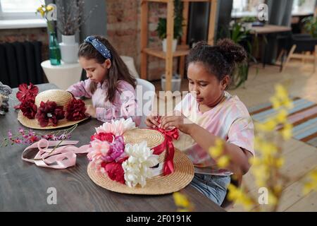 Due bambine interculturali seduti davanti al tavolo macchina fotografica e mettendo nastri di seta e fiori fatti a mano sui cappelli Mentre si prepara per Pasqua Foto Stock