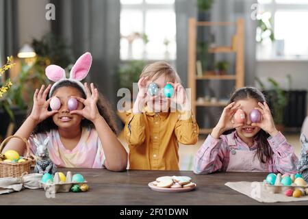 Fila di tre allegri bambini interculturali in casualwear tenuta Dipinse le uova di Pasqua dai loro occhi mentre si siedono da grandi tavolo di legno Foto Stock