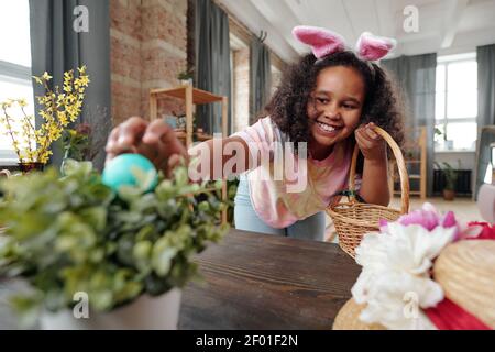 Felice ragazza mista-razza con sorriso toothy che si piega su legno Tavolo mentre mettendo dipinto uovo di Pasqua in vaso con domestico pianta Foto Stock