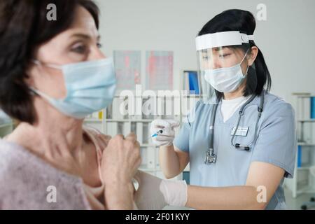Giovane infermiera di etnia asiatica in indumenti da lavoro protettivi che fanno l'iniezione a donna matura seduta davanti a lei in medicina ufficio di cliniche moderne Foto Stock