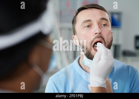 Faccia di giovane paziente maschio che sta provando per la covid con tampone orale tenuto da un'infermiera o da un medico di sesso femminile con guanti seduto di fronte a lei Foto Stock